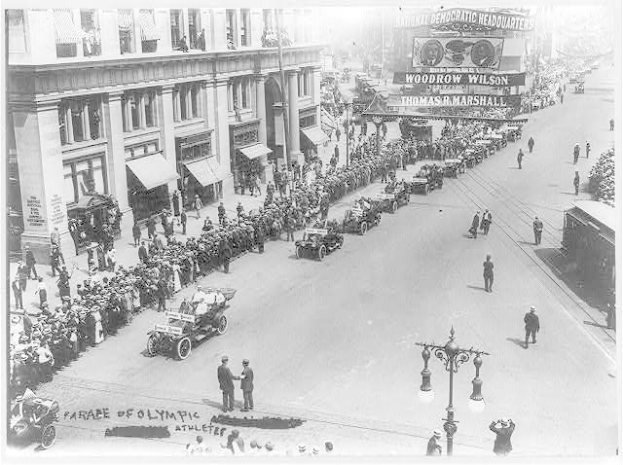 Ein altes Schwarz-Weiß-Foto eines Umzugs auf einer Straße, mit Menschen, Fahrzeugen, Straßenpfählen, einem Schild, Gebäuden mit Fenstern und einem bewölktem Himmel.