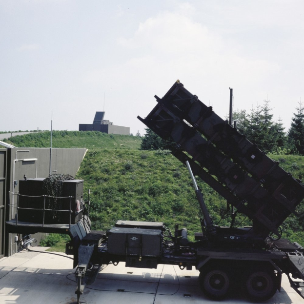 Militärlastwagen mit einer auf der Oberseite montierten Rakete, der vor einem Gebäude mit Bäumen und einem klaren blauen Himmel geparkt ist.