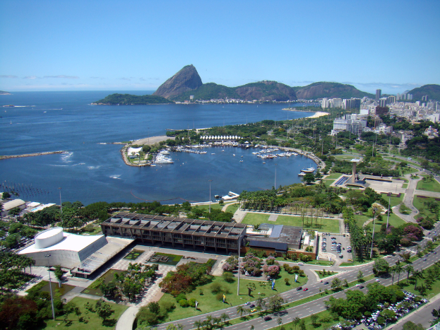Eine Luftaufnahme mit vielen Gebäuden, Straßen, Bäumen, Fahrzeugen, einem Gewässer, Hügeln und dem Himmel.