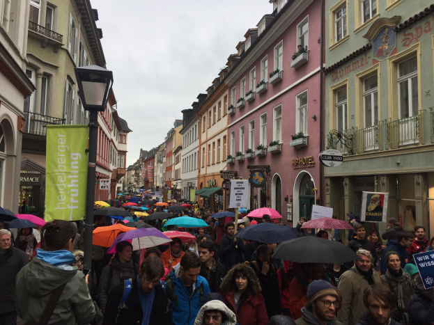 Große Gruppe von Menschen bei einem Klimastreik in Deutschland mit Schirmen, Taschen und Schildern auf der Straße unterwegs, mit Gebäuden und einem Laternenpfahl im Hintergrund.