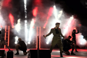 Drake performs auf der Bühne beim Coachella Valley Music and Arts Festival 2018, ein Mikrofon haltend, umgeben von tanzenden Menschen in einer Hockstellung, mit Lautsprechern, einer Kameramann, Metallstangen und einer Decke mit Deckenleuchten.