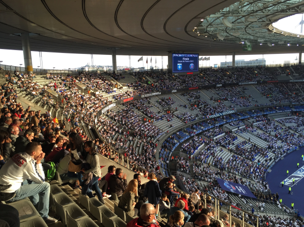 Große Menschenmenge in einem Stadion bei einem Fußballspiel mit einer Bühne, Flaggen, Stangen, einem Bildschirm und der Allianz Arena in München, Deutschland im Hintergrund.