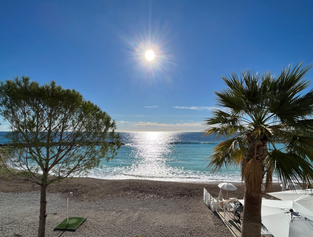 Ein sonniger Strand mit Palmen, Sonnenschirmen und üppiger Vegetation, vor einem hellblauen Himmel mit der Sonne im Hintergrund.