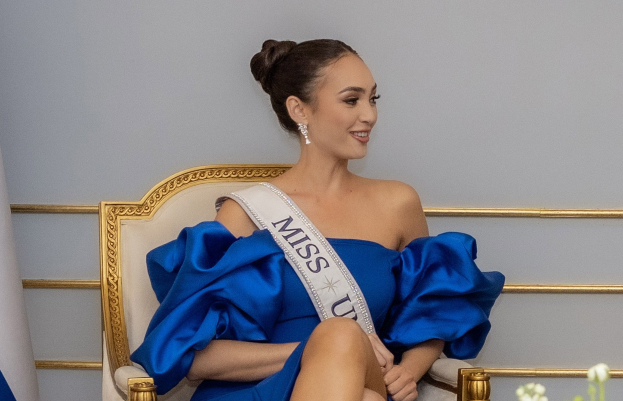 Frau in einem blauen Kleid mit einem "Miss Universe 2019"-Band, die auf einem Stuhl sitzt und lächelt, umgeben von Blumen und einer Wand im Hintergrund.