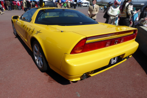 Ein gelber Sportwagen vor einer Menge geparkt, einige Menschen tragen Helme, eine Wand rechts und im Hintergrund Zelte, Bäume, Gebäude und einen klaren blauen Himmel.