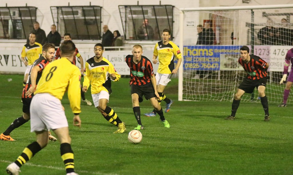 Ein Sportplatz mit mehreren Männern, die Fußball spielen, ein Netz auf der rechten Seite, eine Tafel mit Text im Hintergrund und Zuschauer, die hinter der Tafel stehen, sowie eine Wand und Fenster im Hintergrund.