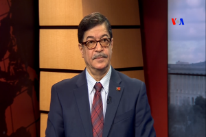 Ein Mann im Anzug und Krawatte, der eine Brille trägt, sitzt ernst vor einem Fernseher.