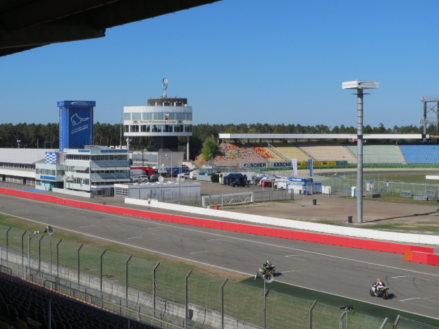Eine Gruppe von Menschen auf Motorrädern auf einer Rennstrecke, umgeben von einem Zaun, Fahrzeugen, einem Gebäude, Schildern, Pfählen, einem Stadion, Lampen, Bäumen und einem bewölkten Himmel.