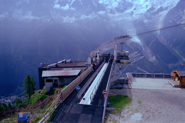 Seilbahn fährt einen schneebedeckten Berg hinauf, mit einem Hintergrund aus schneebedeckten Bergen, die über eine Straße mit Schildern, Geländern, Vegetation und Häusern fahren, mit einigen wenigen Menschen sichtbar.