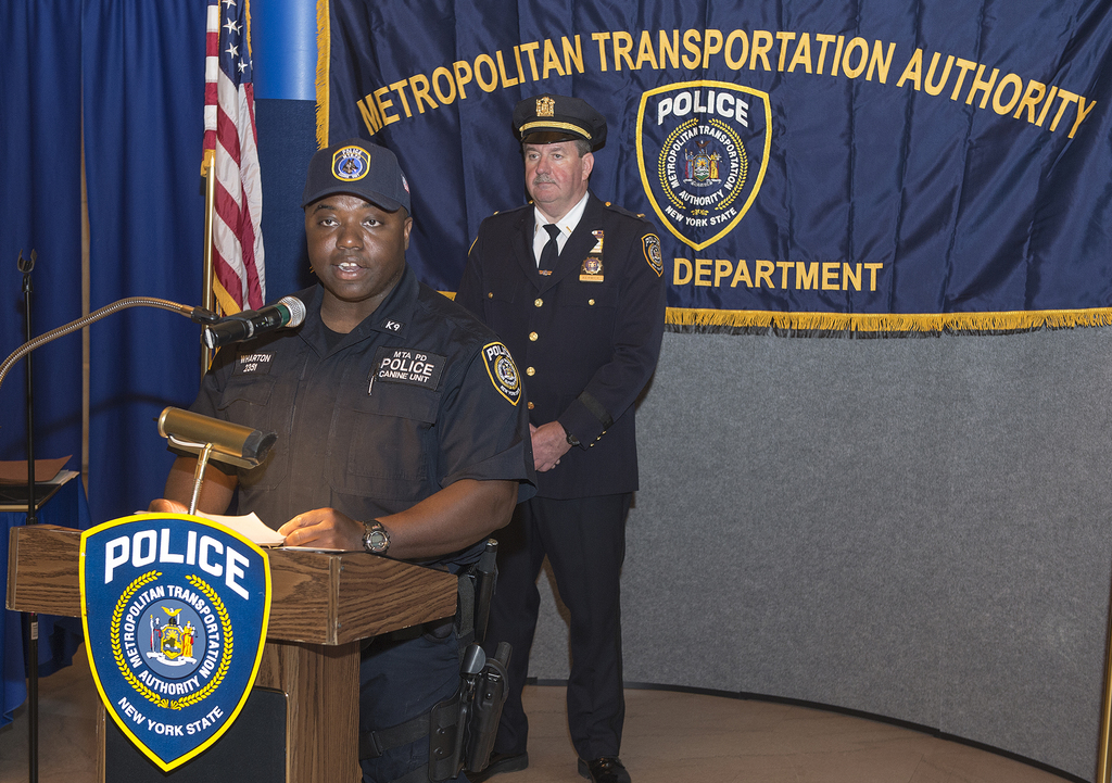 Zwei Polizeibeamte stehen vor einem Rednerpult, einer hält Papiere und spricht in ein Mikrofon, mit einer Fahne, einem Banner und einem Tisch in der Nähe und einer Tafel mit Symbolen und Text im Hintergrund.