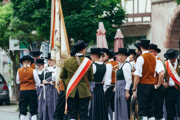 Gruppe von Menschen in traditioneller bayrischer Kleidung, die eine Straße entlangmarschieren, einige halten Musikinstrumente und Fahnen, mit Bäumen, Gebäuden und einem Auto im Hintergrund.