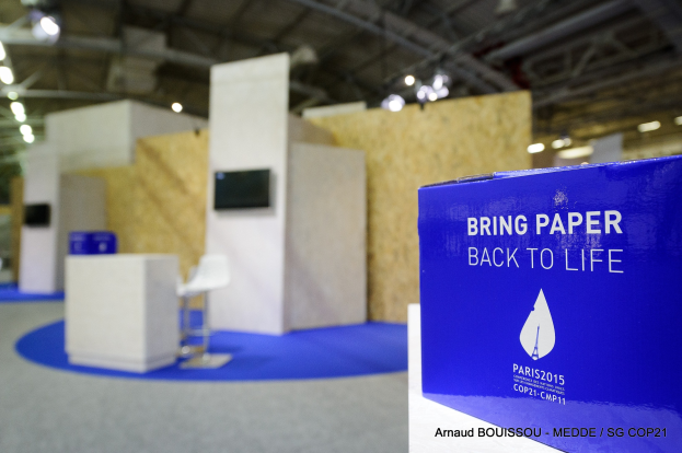Ein Messestand mit einem blauen und weißen Schild, auf dem "Bring Paper Back to Life" steht, auf der rechten Seite, mit unscharfen Stühlen, Tischen, einem wandmontierten Fernseher und Deckenlampen im Hintergrund.