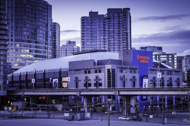 Eine Stadtansicht mit hohen Gebäuden, einer Brücke mit Säulen und Bögen, Fahrzeugen auf der Straße, Straßenlaternen, Schildern und einem bewölkten Himmel, identifiziert als Rogers Arena in Toronto, Ontario.