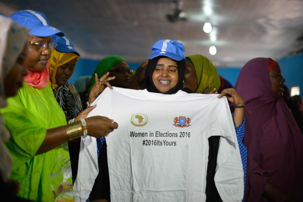 Eine Gruppe von Frauen, die ein weißes T-Shirt mit der Aufschrift "Women in Elections 2016" hochhalten, einige tragen blaue Mützen, unter Deckenbeleuchtung.