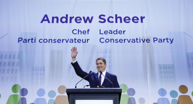 Konservative-Parteichef Andrew Scheer steht vor einem Podium mit zwei Mikrofonen und einem Bildschirm dahinter, vor einem Banner mit Text und Gebäudeillustrationen.