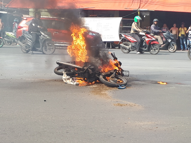 Motorrad in Flammen am Straßenrand mit einem Auto und einem Van im Hintergrund, umgeben von Menschen, einige tragen Helme, mit einem Banner und einem Zelt im Bild.