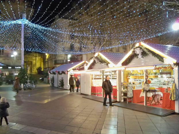 Ein lebendiger Weihnachtsmarkt bei Nacht in einer Stadt, mit Menschen, die stehen, sitzen und ein Kind halten, umgeben von festlichen Lichtern, Pfählen, Pflanzen, Bäumen und Gebäuden mit Fenstern unter einem bewölkten Himmel.