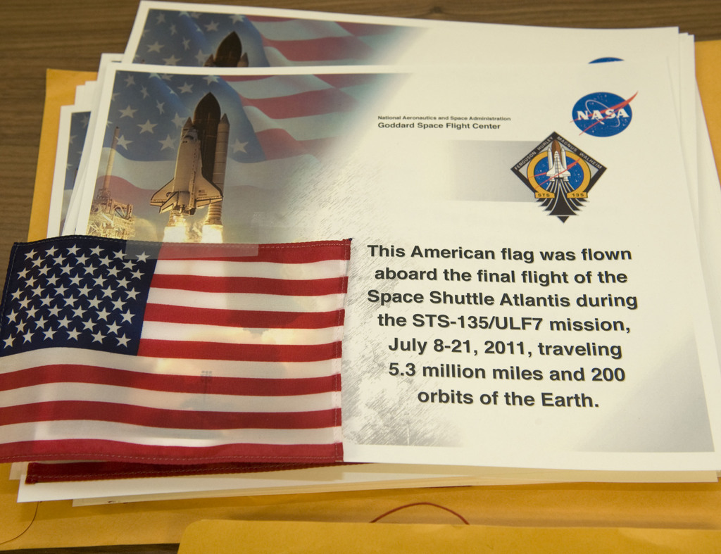 Ein zentrales Element sind Plakate mit Text, einer Flagge, einer Rakete und verschiedenen Logos, mit einem Tisch unten und einem Boden oben im Bild.