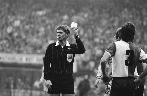 Ein Schiedsrichter in einem Schwarz-Weiß-Bild hält eine rote Karte vor einer Menge von Menschen in Fußballuniformen hoch, mit einem Zaun im Hintergrund.