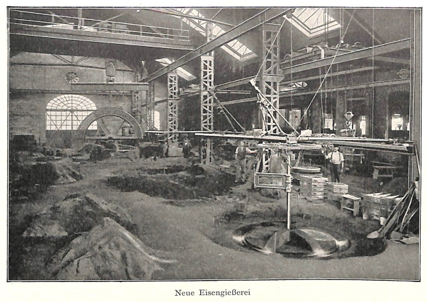 Altes Schwarz-Weiß-Foto einer Fabrik mit Arbeitern, die an einer Maschine arbeiten, umgeben von Metallstangen und -rohren, beschriftet mit "Neue Eisenglebeerei, die erste Fabrik Deutschlands" unten.