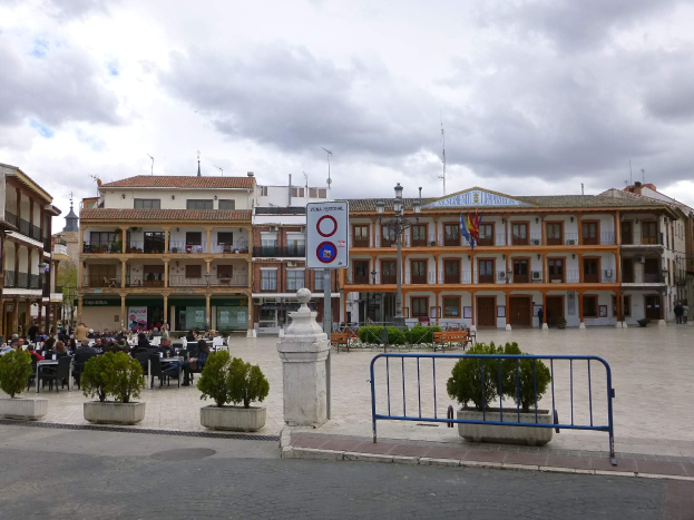 Ein belebter Stadtplatz mit Menschen, die sitzen und stehen, umgeben von Topfpflanzen, Metallabsperrungen, Schildern, Straßenlaternen mit Fahnen, Gebäuden und einem bewölkten Himmel.