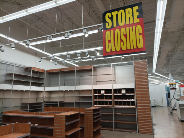 Store interior with a "Store Closing" sign hanging from the ceiling, shelves stocked with items, a board with text, and overhead lighting.