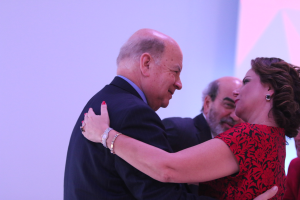 Ein Mann und eine Frau umarmen sich herzlich auf einer Bühne, umgeben von ein paar anderen Menschen; der Mann trägt ein Armband.