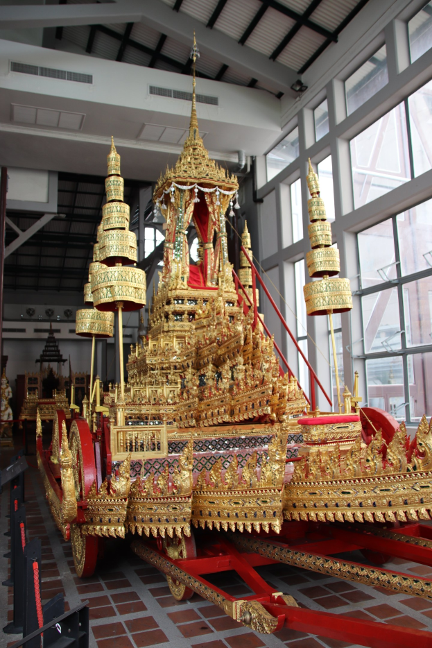 Ein großer goldener Wagen mit kunstvollen Verzierungen befindet sich in der Mitte eines Raumes mit Glasfenstern auf der rechten Seite und einer Decke darüber.