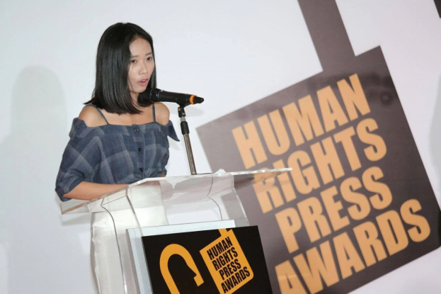 Frau in schwarzem Kleid und Dutt steht an einem Rednerpult mit Mikrofon und hält eine Rede vor einem Banner 'Human Rights Press Awards'.