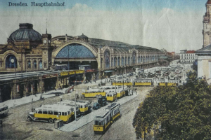 Ein altes Postkartenbild zeigt einen belebten Dresdner Bahnhof mit Fahrzeugen, Menschen, Bäumen, Gebäuden und einem bewölkten Himmel, mit der Aufschrift «Dresden Hauptbahnhof» oben.