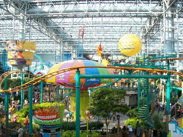 Ein großer Indoor-Wasserpark mit einer Achterbahn, die von Menschen, Bäumen, Pflanzen und Deckenbeleuchtung umgeben ist.