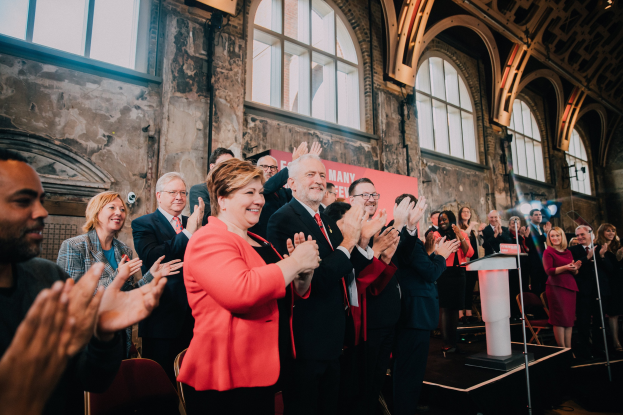 Menschen in einem Raum klatschen, mit einem Podium, einem Mikrofon, Stühlen, einer Tafel mit der Aufschrift "Covid-19 New York City Mayor Bill de Blasio", einer Wand und Fenstern im Hintergrund.