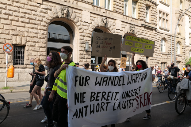 Eine Gruppe von Menschen marschiert auf einer Demonstration gegen die Anti-Kriegsbewegung in Berlin, einige halten Schilder und andere fahren Fahrräder, im Hintergrund ein Gebäude mit Bögen, Säulen und Skulpturen.