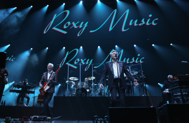Eine Gruppe von Musikern, die auf der Bühne mit Instrumenten, Mikrofonen und Ständern spielen, mit Kabeln und Equipment im Vordergrund und einem Schild im Hintergrund mit der Aufschrift "Roxy Music - Live in der O2 Arena, London".