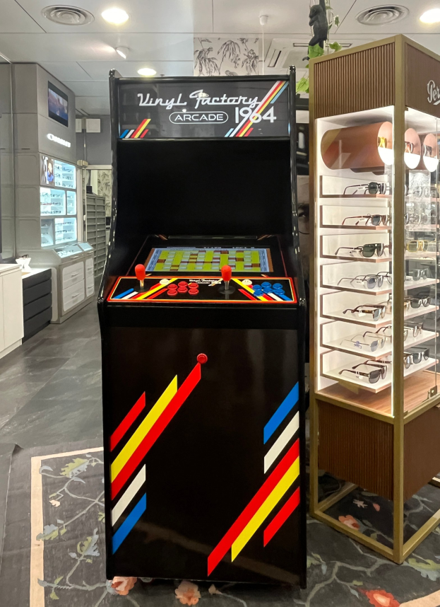 Ein Geschäftsinnenraum mit einem zentralen Retro-Arcade-Automaten, einer Glasvitrine auf der rechten Seite und einer retro-stilvollen Beleuchtung mit Schränken und einem Fernseher im Hintergrund.