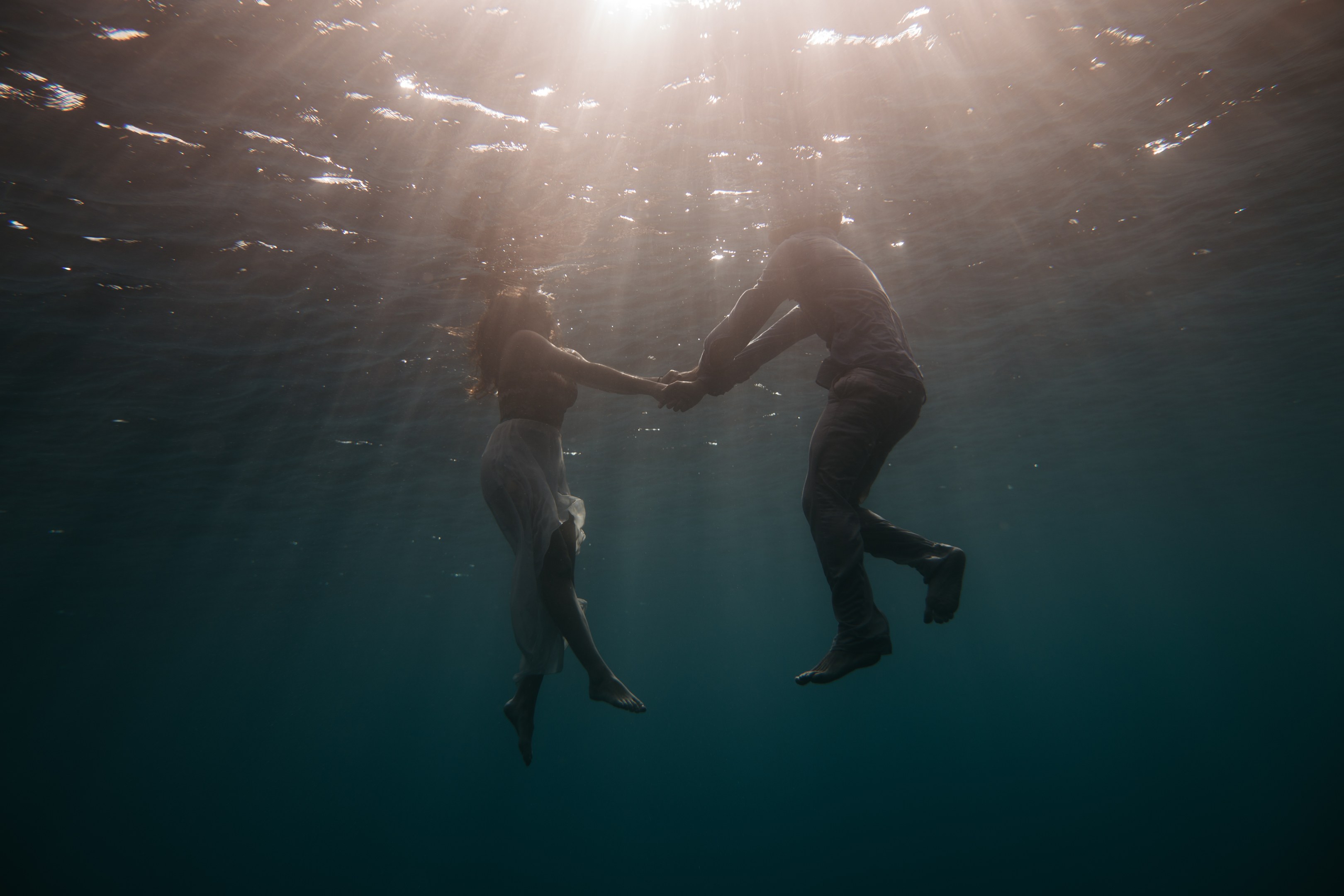 Zwei Personen, die sich unter Wasser in der See an den Händen halten, mit Sonnenstrahlen, die von oben scheinen.