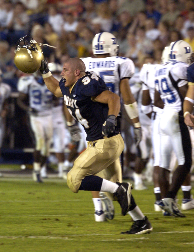 Ein Fußballspieler in einem weißen Trikot rennt mit dem Ball, umgeben von Teamkollegen in Sportkleidung und Helmen auf dem Feld, während Zuschauer auf Stühlen im Hintergrund sitzen.