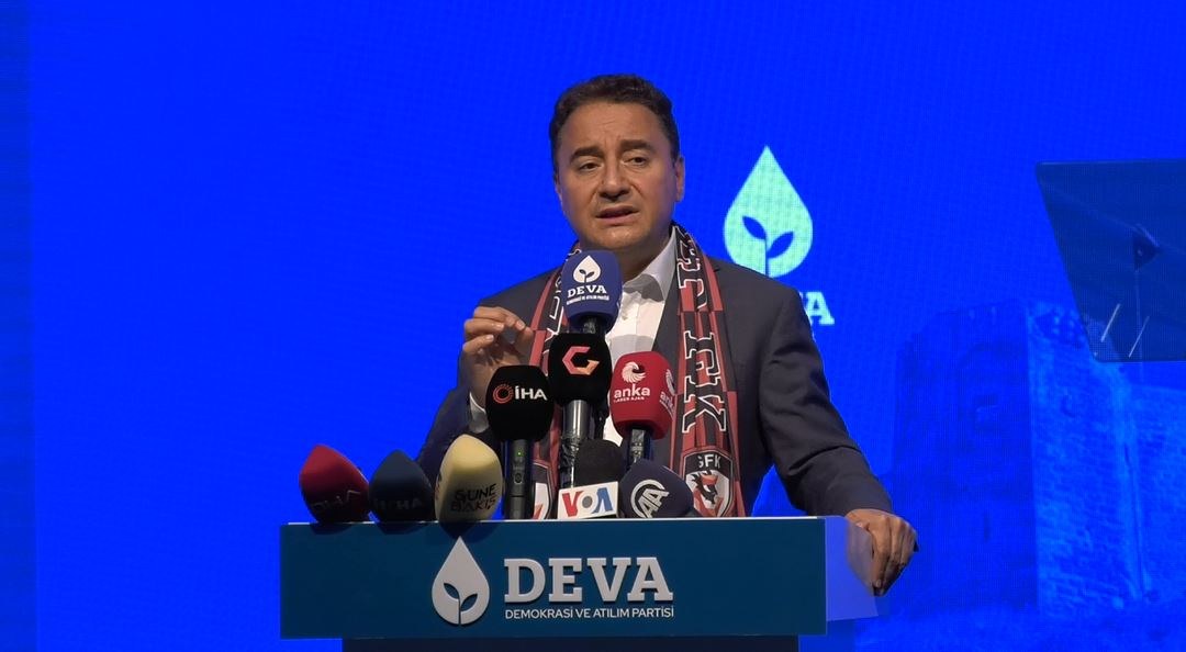 Ein Mann in Mantel und Schal steht hinter Mikrofonen auf einem Podium und spricht vor einer Tafel mit der Aufschrift "deva-Gesch├Ąftsführer sagt, es sei keine gute Idee für die Zukunft.", mit einem Bildschirm und einem Objekt im Hintergrund.