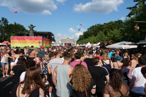 Eine große Menschenmenge, die eine Straße mit Zelten, Bäumen, Pfählen und Lichtern entlanggeht, mit Gebäuden und einem bewölkten Himmel mit Ballons im Hintergrund beim Berliner Christopher Street Day.