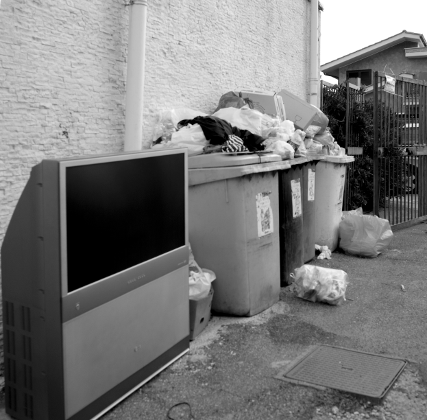 Eine Schwarz-Weiß-Szene eines weggeworfenen Fernsehers auf der Straße neben einem Haufen Müll, mit vollen Mülltonnen und anderen Gegenständen im Vordergrund und einer Wand, einem Rohr, einem Tor, Pflanzen, einem Auto und einem Gebäude im Hintergrund.