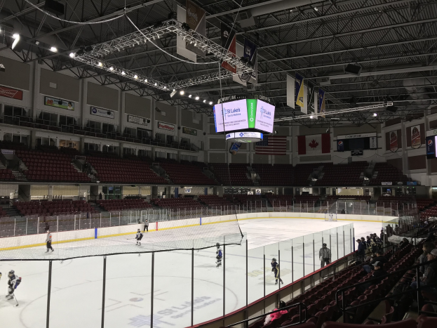 Ein Hockey-Spiel in einer großen Arena mit einem zentralen Netz, Zuschauern auf beiden Seiten, Deckenleuchten und Trägern sowie Bannern und Fahnen an den Wänden, identifiziert als die St. Louis Blues Arena.