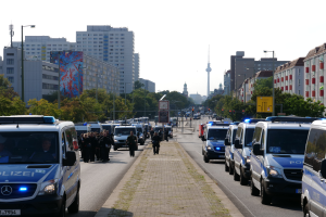 Eine große Gruppe von Polizeiwagen parkt am Straßenrand in Berlin, Deutschland, mit vielen Menschen auf dem Gehweg, Bäumen, Gebäuden, Laternen, Verkehrszeichen, Schildern mit Text und einem Turm im Hintergrund.
