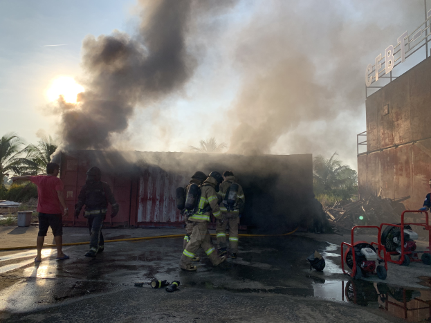 Eine Gruppe von Feuerwehrleuten in Helmen steht vor einem brennenden Gebäude mit aufsteigendem Rauch, während ein Gebäude mit einer Namensplakette auf der rechten Seite und Bäume auf der linken Seite sichtbar sind.