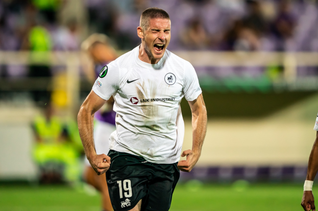 Ein Mann in weißem Hemd und schwarzen Shorts feiert auf einem Fußballfeld umgeben von einer Gruppe von Menschen, wahrscheinlich nach einem Tor in einem Spiel zwischen Leeds United und Melbourne Victory.