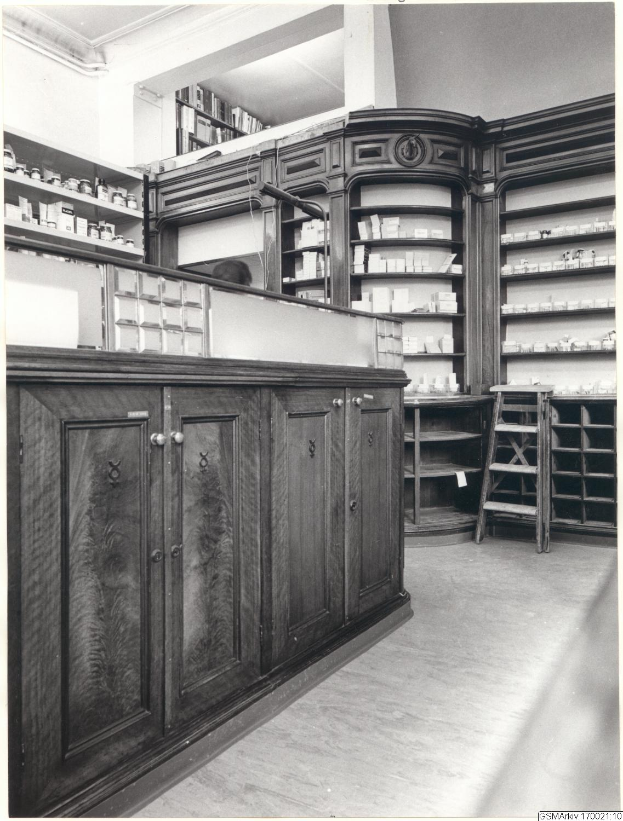 Ein Schwarz-Wei├č-Foto einer Apotheken-Inneneinrichtung mit einer Theke, Sch├╝ben, einer Leiter und Regalen, die mit verschiedenen Gegenst├Ąnden gef├╝llt sind, mit Text ├╝ber dem Bild.