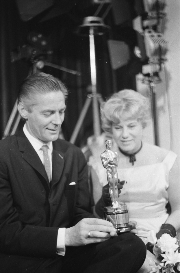 Ein Mann in einem schwarzen Blazer, weißem Hemd und Krawatte hölt eine Oscar-Statue, waährend er neben einer Frau in Abendgarderobe sitzt, die einen Blumenstrauß hölt, beide in Schwarz-Weiß.