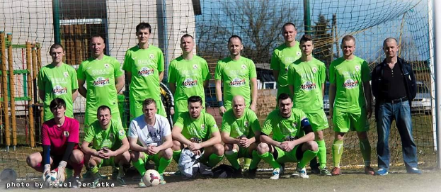 Gruppe von Männern in grünen T-Shirts, die auf einem Füßballfeld in der Nähe eines Torpfostens stehen oder hocken, mit einem Ball vor ihnen und verschiedenen Hintergrundelementen, darunter Bäume, Gebäude und Fahrzeuge.