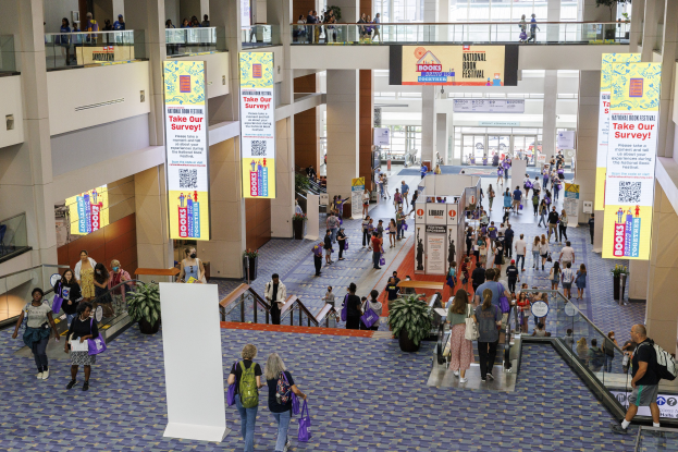 Große Gruppe von Menschen, die durch ein großes Gebäude mit Bannern, Topfpflanzen, Rolltreppen, Geländern, Säulen, bedruckten Tafeln, Glastüren und anderen Objekten gehen, einige tragen Taschen, was auf den Besuch der Nationalen Buchmesse hinweist.