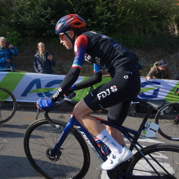 Professionelle Radrennfahrerin Katie Archibald mit Helm, Schutzbrille, Handschuhen und Schuhen auf einem Fahrrad die Straße entlang fahrend, vorbei an einer Zuschauermenge, mit einem Banner, einer Wand, Bäumen und einem Gebäude im Hintergrund.