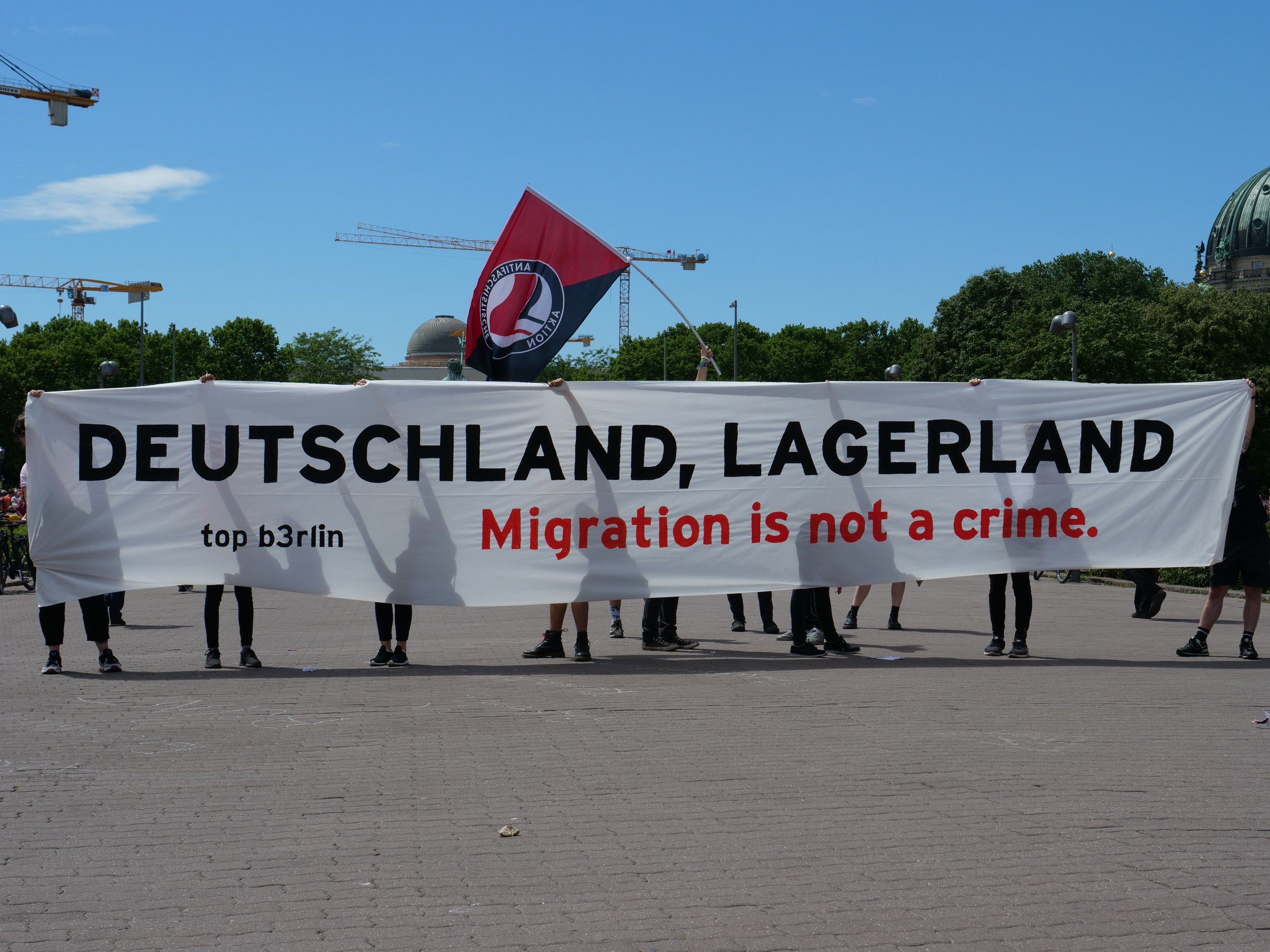 Eine Gruppe von Menschen mit einem Banner "Deutschland, Lagerland Migration ist kein Verbrechen" und einer Flagge, mit Straßenlaternen, Bäumen, Kränen, einem Gebäude und einem bewölkten Himmel im Hintergrund.